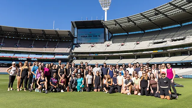 Large group photo on the MCG turf with people in activewear. Sign says Yoga on the 'G.