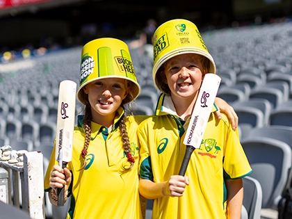 Two young cricket fans wearing Australian cricket gear, bucket hats and holding small cricket bats at the MCG