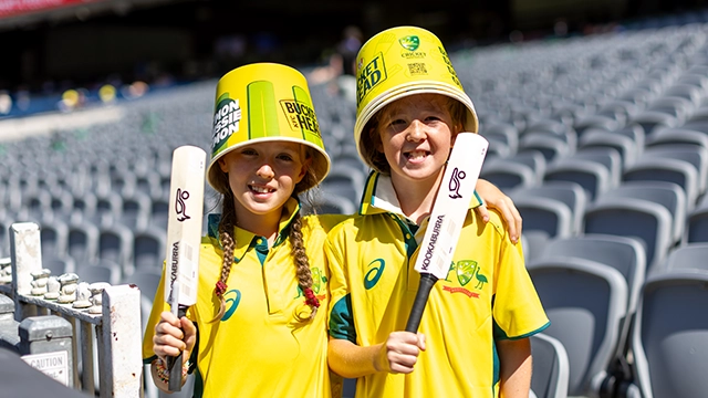 Two young cricket fans wearing Australian cricket gear, bucket hats and holding small cricket bats at the MCG