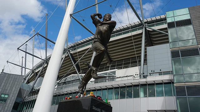 Bronze statue of Shane Warne bowling, on a backdrop of the MCG exterior.