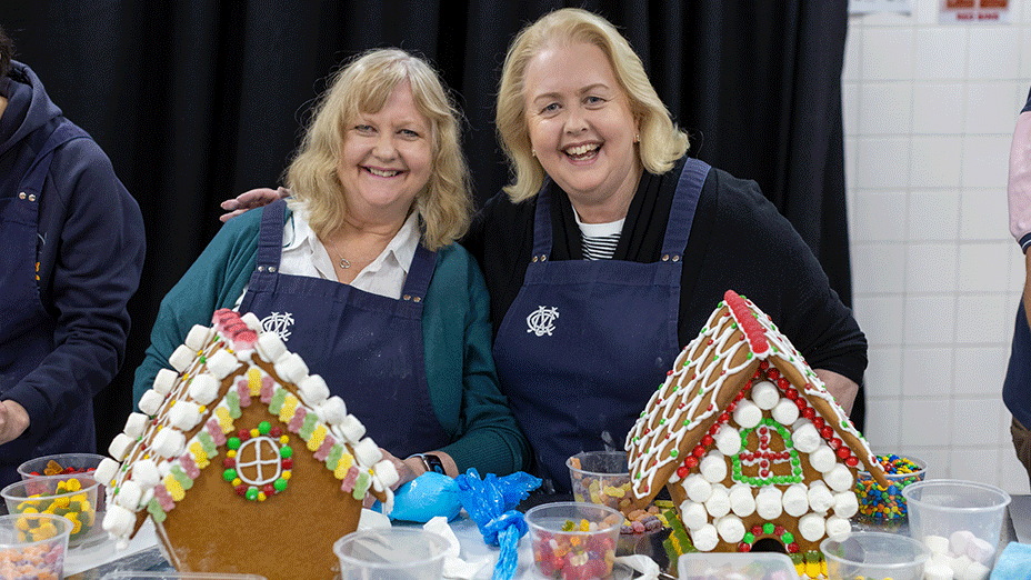 Two participants in MCC aprons smiling with their decorated gingerbread houses